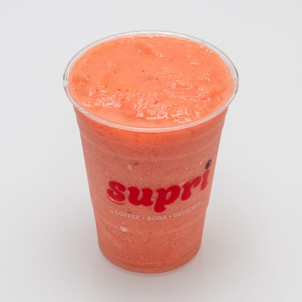 A vibrant pink-orange Supri smoothie in a clear plastic cup, filled to the brim with a frothy texture.